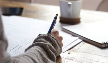 person writing on brown wooden table near white ceramic mug